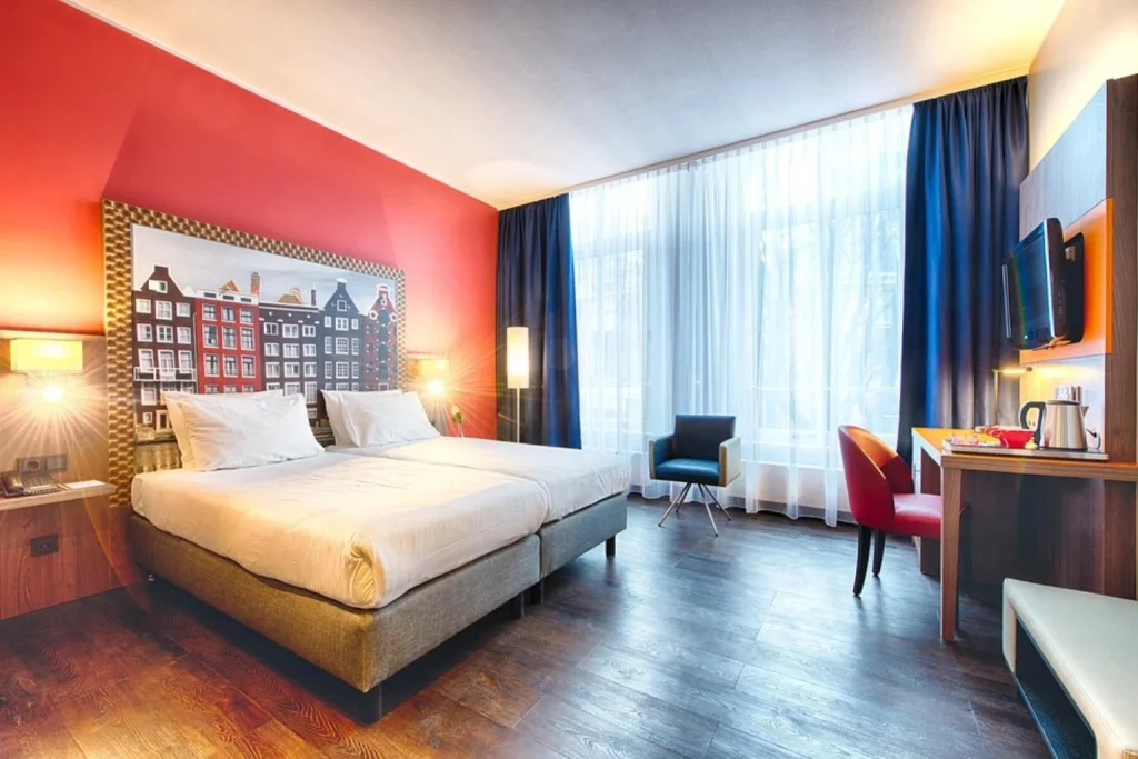 Bright modern guest room with twin beds, red accent wall and large windows at Leonardo Hotel Amsterdam City Center, Amsterdam