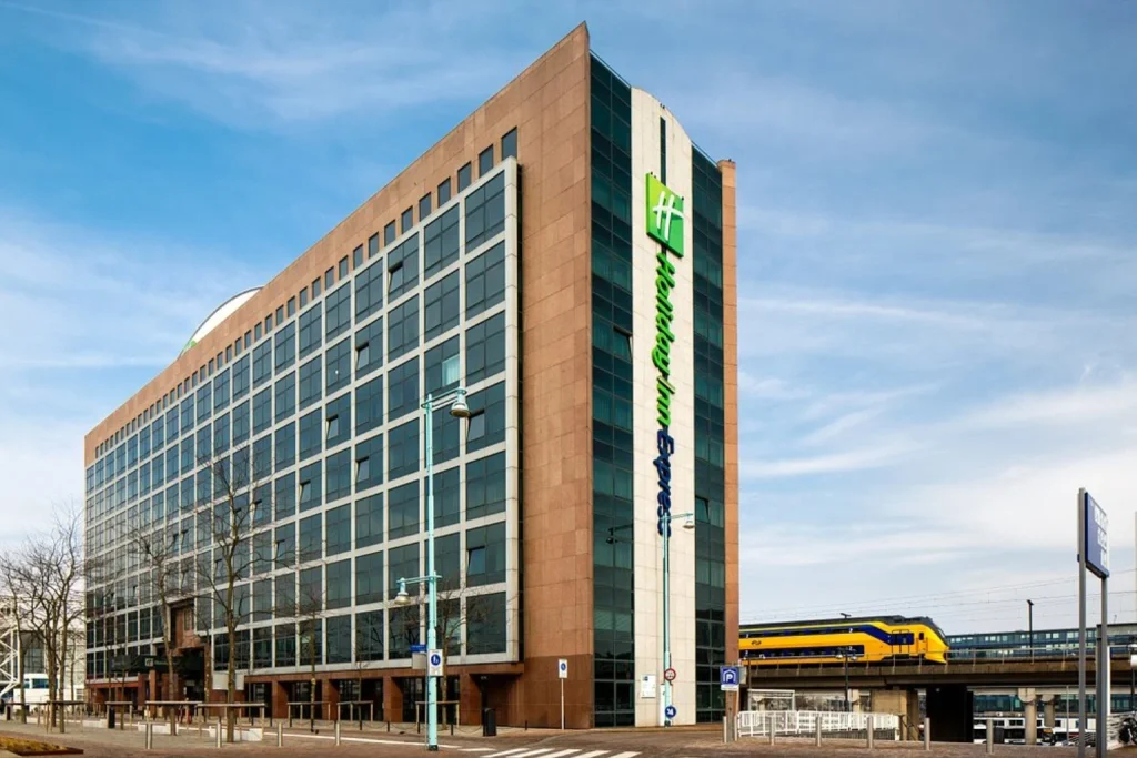 Modern high-rise hotel building next to Sloterdijk Station with a Dutch train passing alongside, Holiday Inn Express Amsterdam – Sloterdijk Station, Amsterdam.