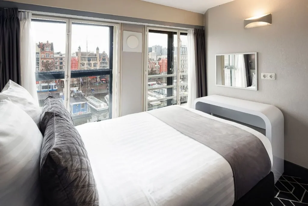Bright double room at Hotel CC featuring a comfortable bed, modern furnishings, large windows, and views of Amsterdam’s canal and surrounding historic buildings.