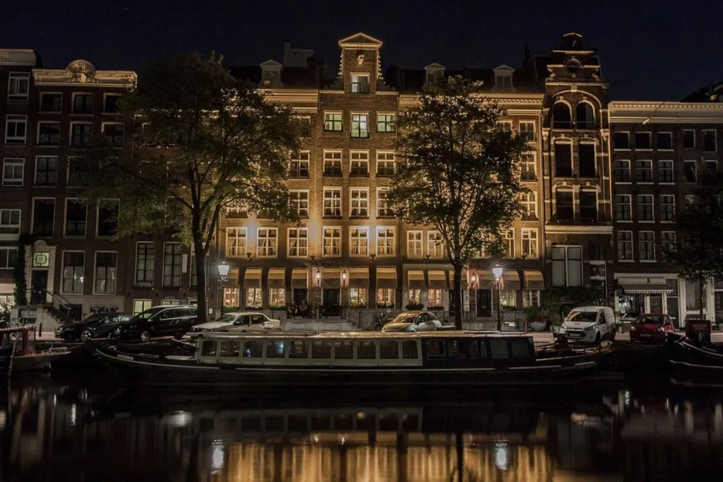 Illuminated historic canal-side façade with warm lights reflecting in the water at night, Hotel Estheréa, Amsterdam.