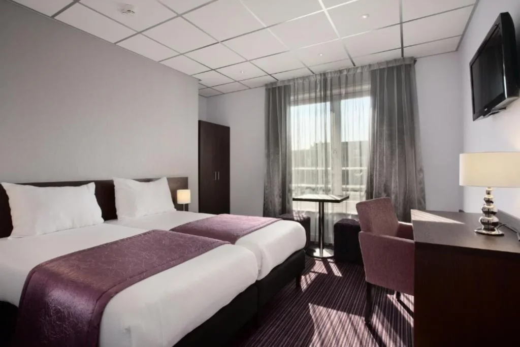 Bright twin-bed room at Hotel Luxer featuring two beds, a seating area, work desk, television, wardrobe, and large windows overlooking the surrounding area.