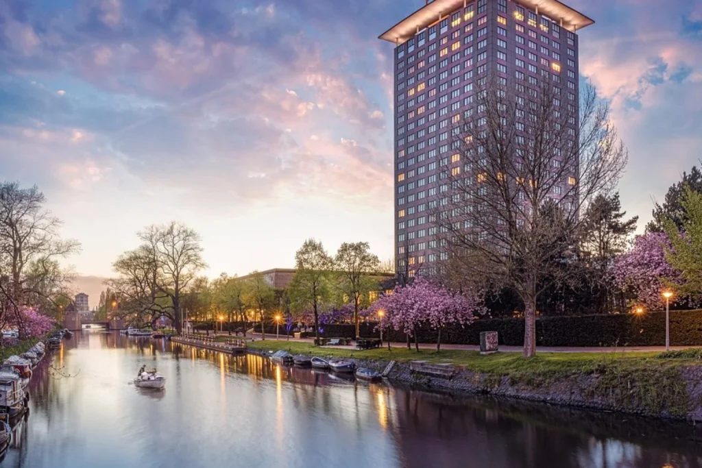 Tall hotel tower beside a canal with flowering trees at sunset, Hotel Okura Amsterdam, Amsterdam