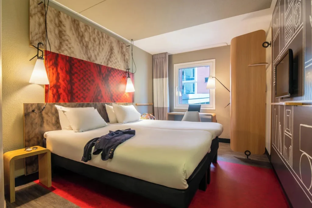 Modern twin room at ibis Amsterdam Centre Stopera featuring two single beds, a compact workspace and colorful contemporary décor.