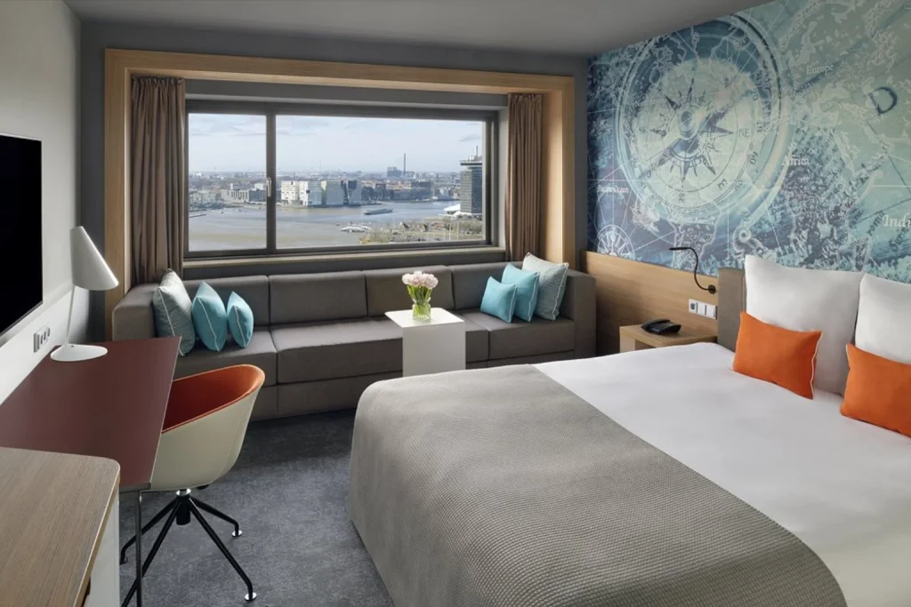 Modern guest room with large window, seating area and panoramic river views at Mövenpick Hotel Amsterdam City Centre, Amsterdam