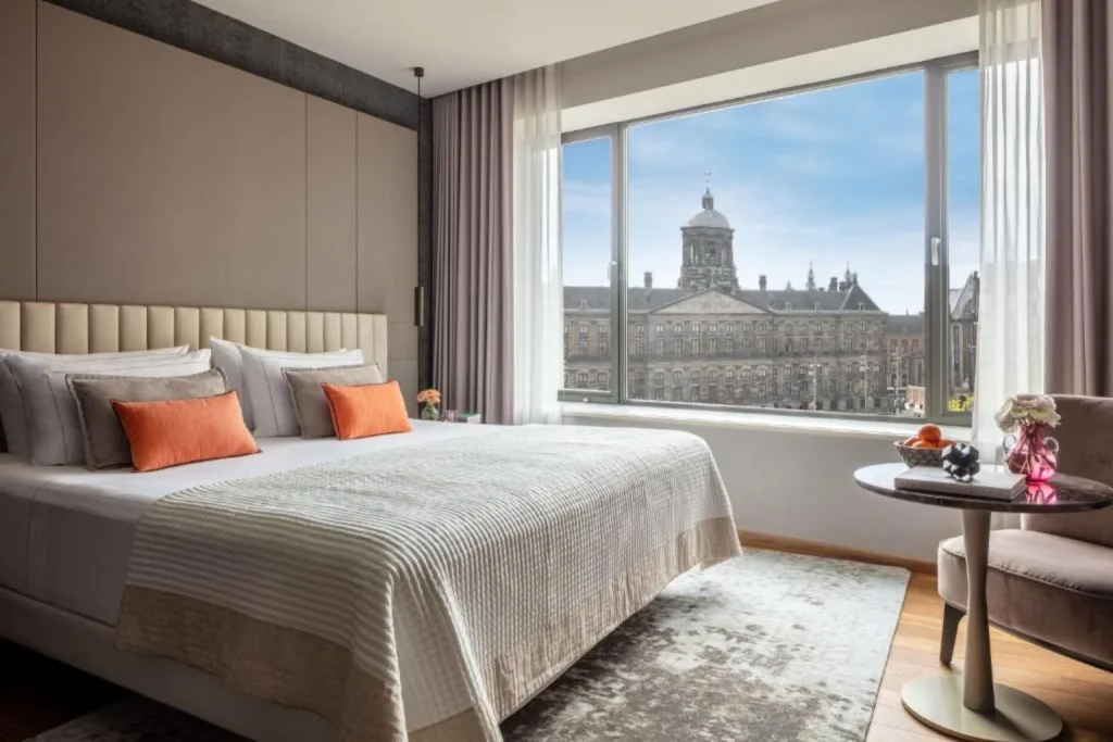 Elegant hotel room featuring a king-size bed, contemporary furnishings, a seating area, and a large window overlooking the Royal Palace in Amsterdam.