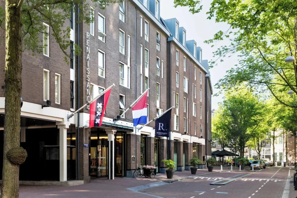 Modern brick facade with Dutch and Amsterdam flags along a leafy city street at Renaissance Amsterdam Hotel.