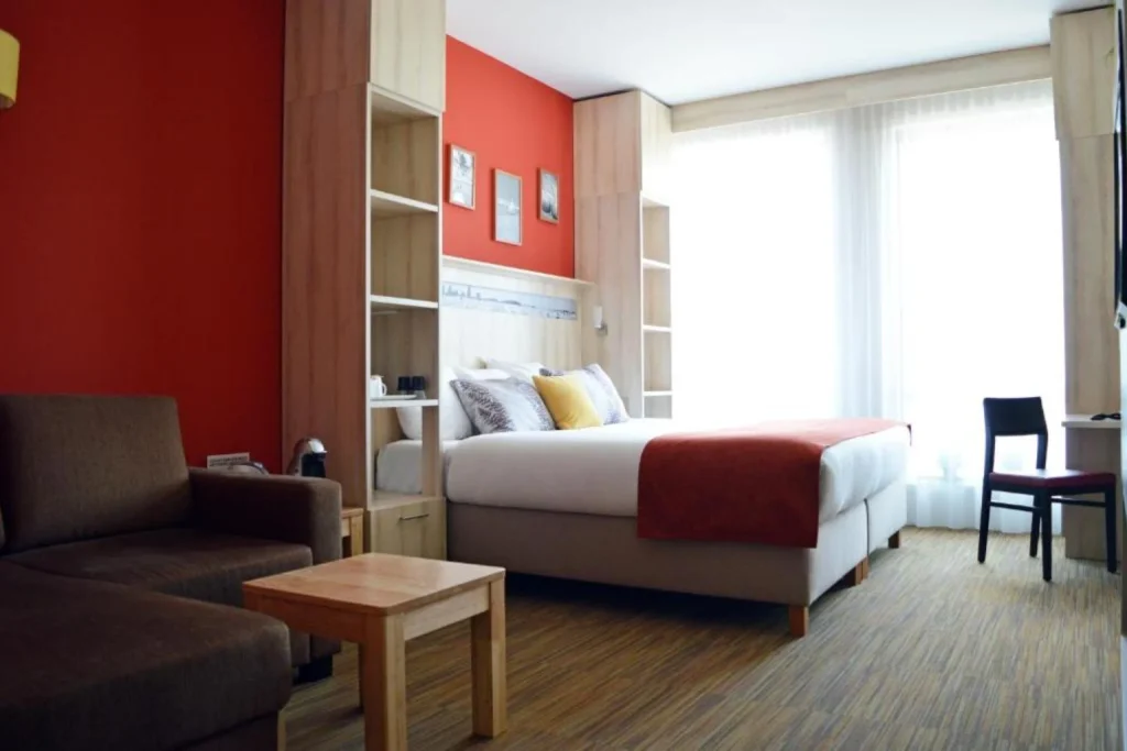 Bright modern guest room with a double bed, warm red accents, wooden furnishings and a seating area at Skotel Amsterdam Hotelschool The Hague, Amsterdam.
