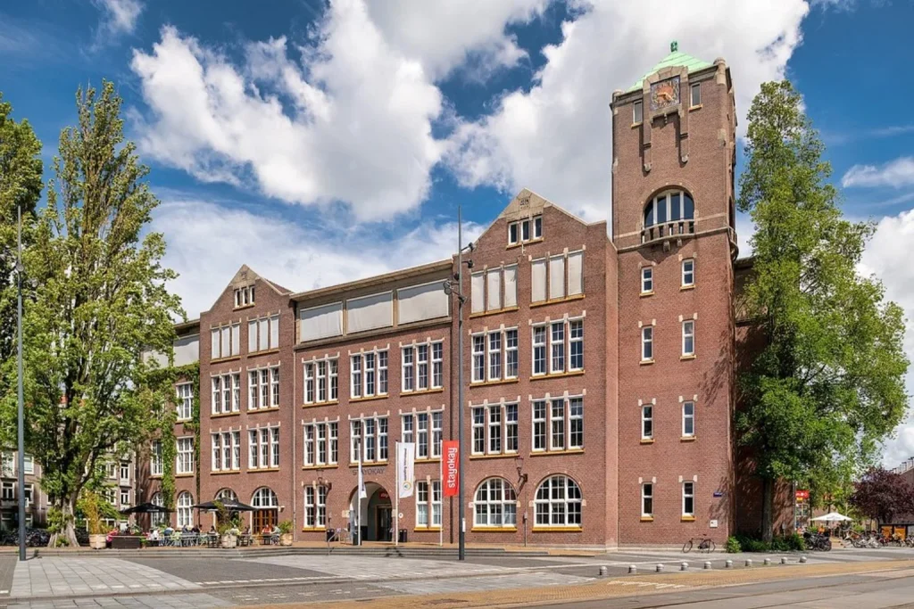 Historic red-brick building with a distinctive clock tower and main entrance, surrounded by mature trees at Stayokay Hostel Amsterdam Oost, Amsterdam.