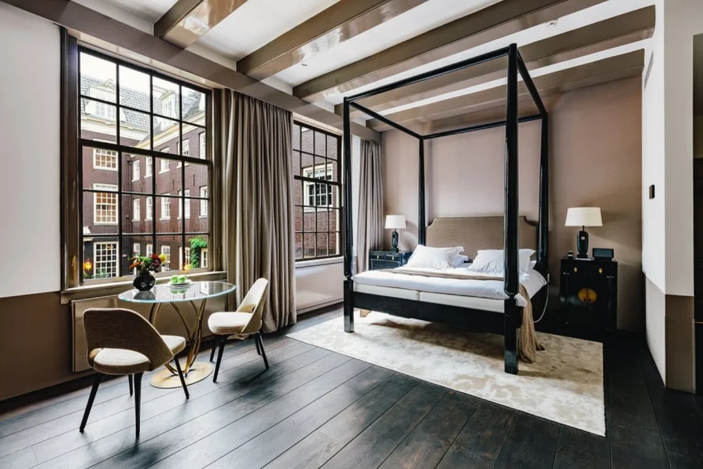 Elegant bedroom featuring a four-poster bed, dark wooden floors, exposed ceiling beams and large historic windows at The Dylan Amsterdam, Amsterdam.