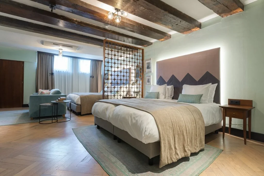 Spacious guest room at The Hendrick’s Hotel Amsterdam featuring multiple beds, exposed wooden ceiling beams, a seating area and distinctive boutique-style décor.