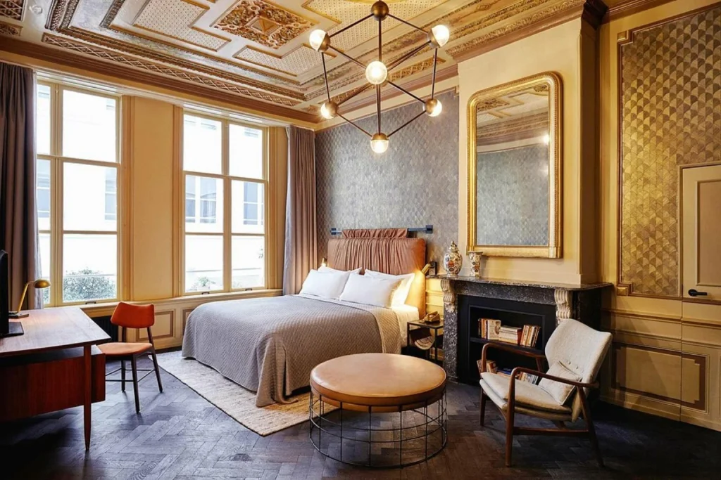 Stylish boutique bedroom featuring an ornate historic ceiling, decorative fireplace, contemporary furnishings and large windows at The Hoxton, Amsterdam.
