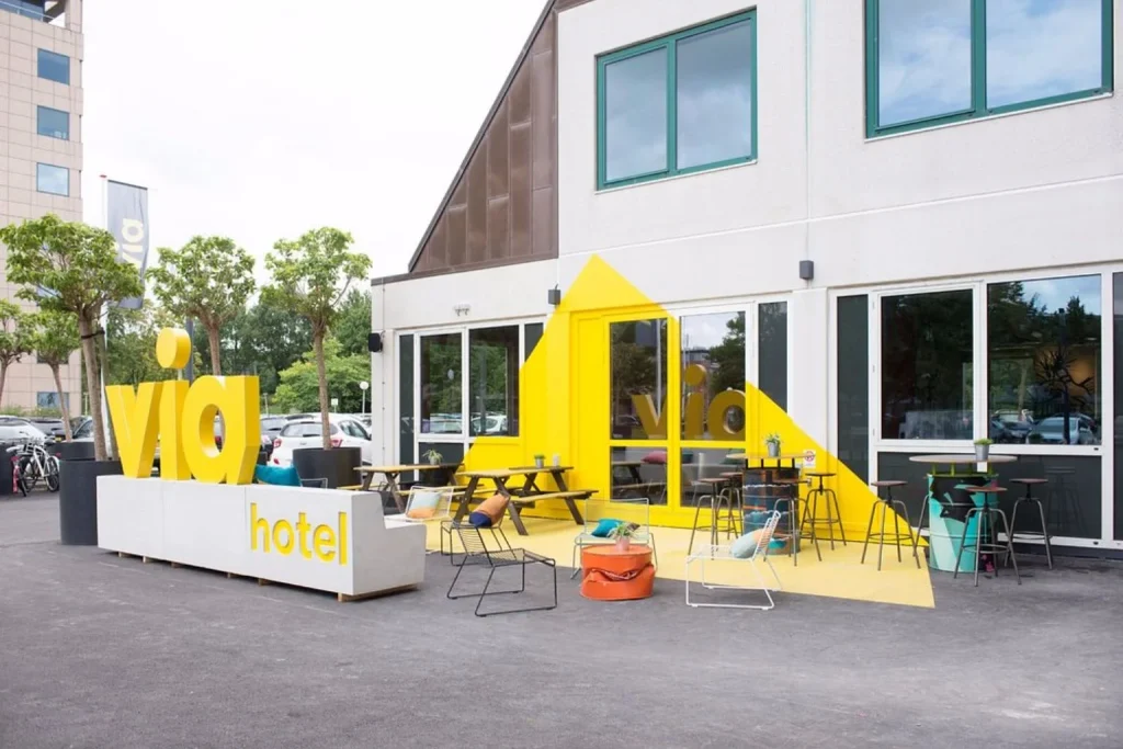 bright and colorful exterior with its distinctive yellow branding, outdoor seating area, and modern communal spaces at Via Amsterdam Hostel, Amsterdam