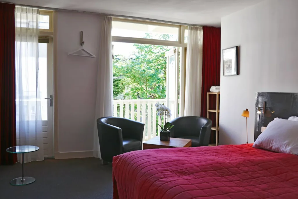 Spacious and bright guest room with a red bed, comfortable seating and balcony overlooking greenery at Adriaen van Ostade B&B, Amsterdam