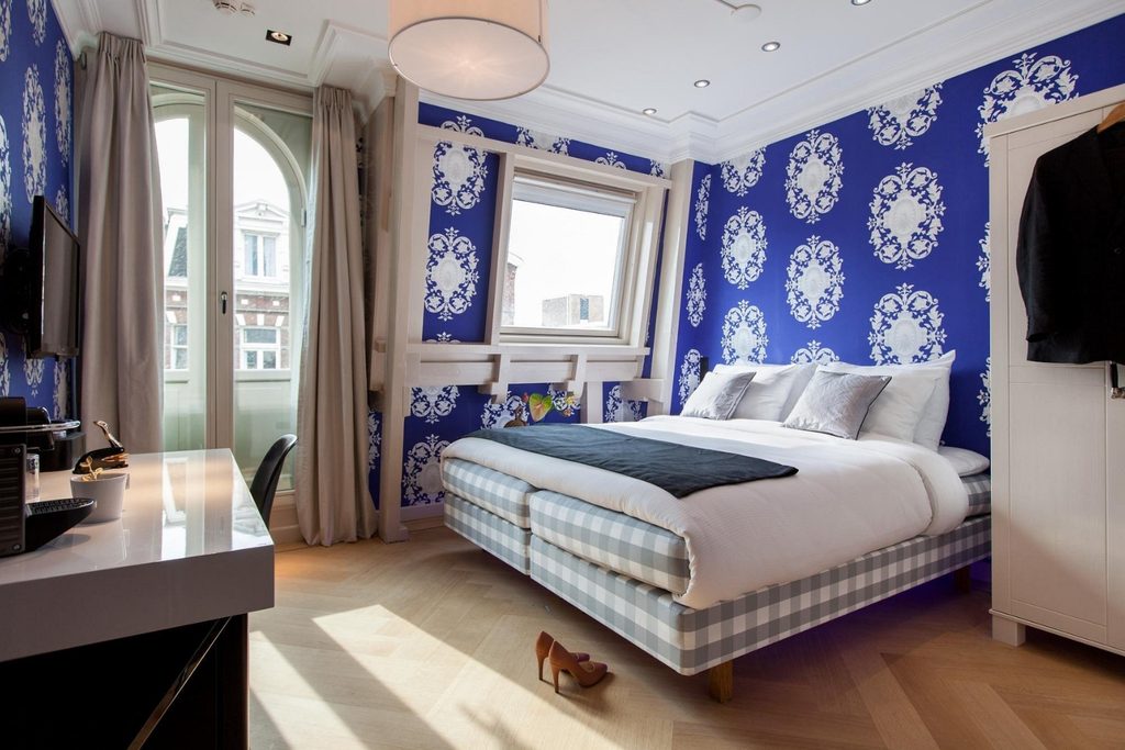 Stylish double room featuring bold blue patterned wallpaper, a checkered bed, large windows, wooden floors and a compact work desk at Amsterdam Canal Hotel, Amsterdam