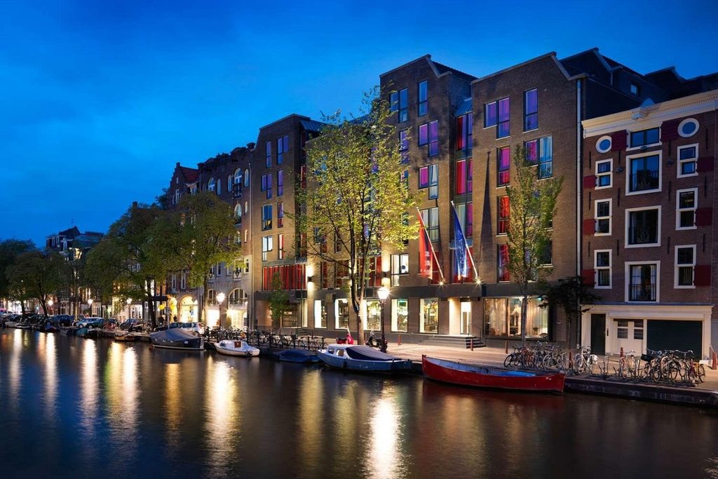 Evening canal view of Andaz Amsterdam Prinsengracht, with illuminated waterfront buildings and boats moored along the canal.