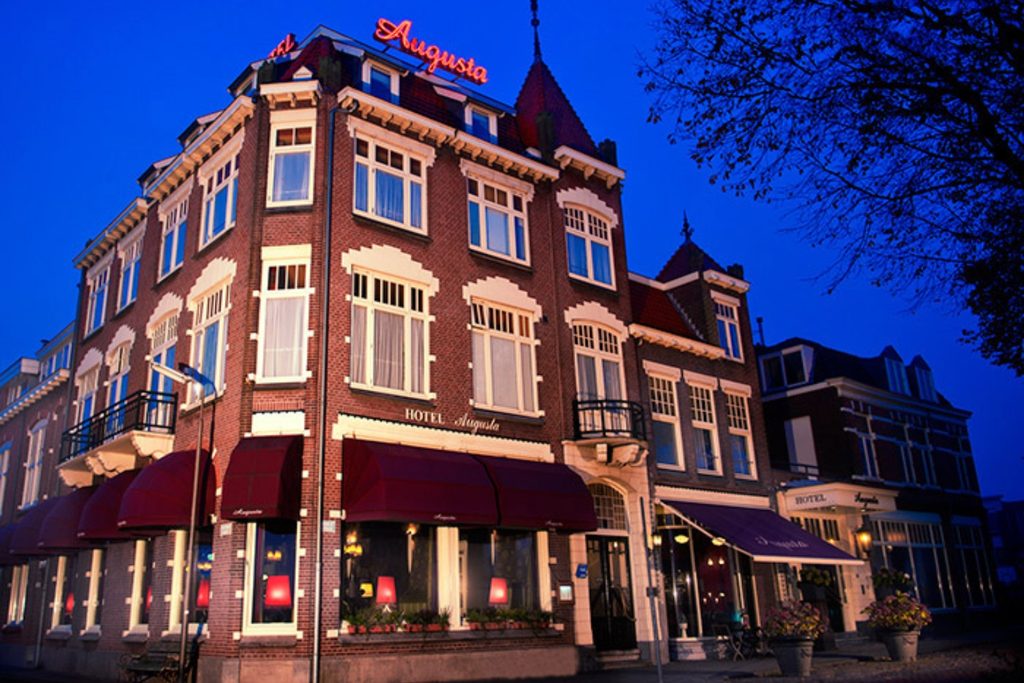 Historic brick façade of Augusta Hotel IJmuiden illuminated at dusk, featuring traditional architecture, large windows and the hotel entrance.