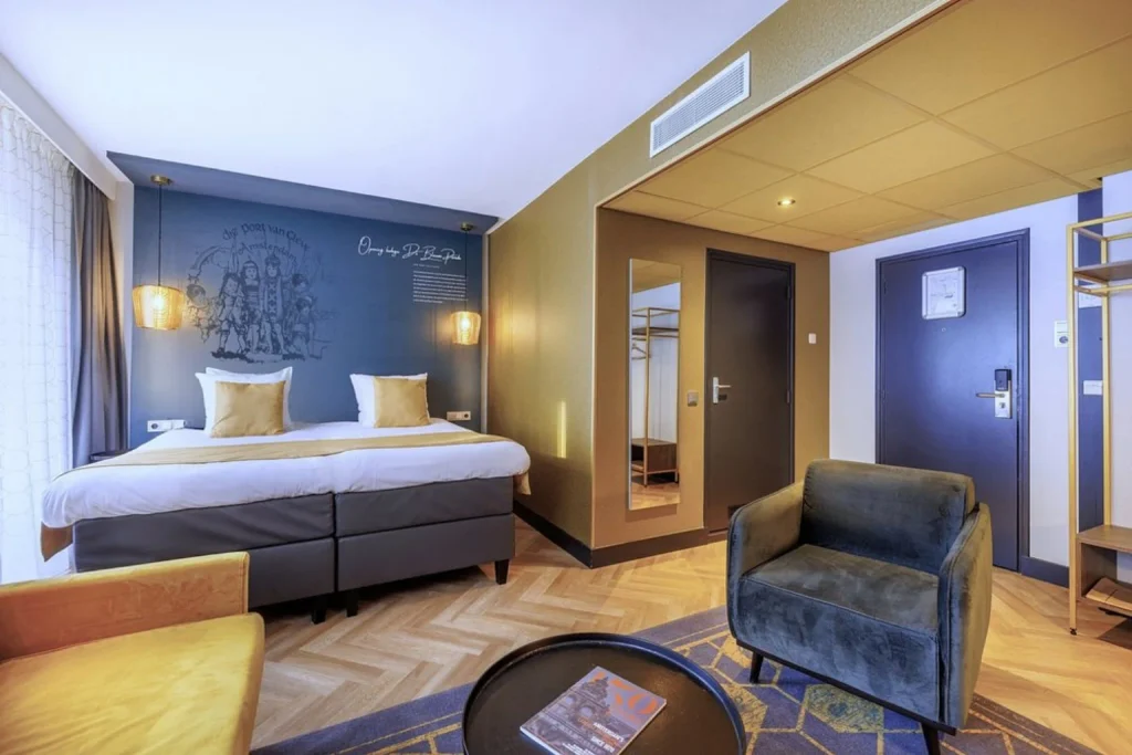 Spacious double room with contemporary furnishings, twin beds, armchairs and warm gold accents at Die Port Van Cleve Amsterdam.