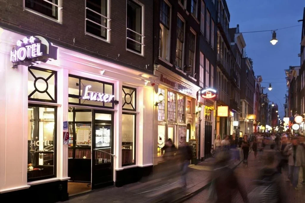 Illuminated hotel entrance surrounded by historic buildings and a lively city street in the evening at Hotel Luxer Amsterdam.