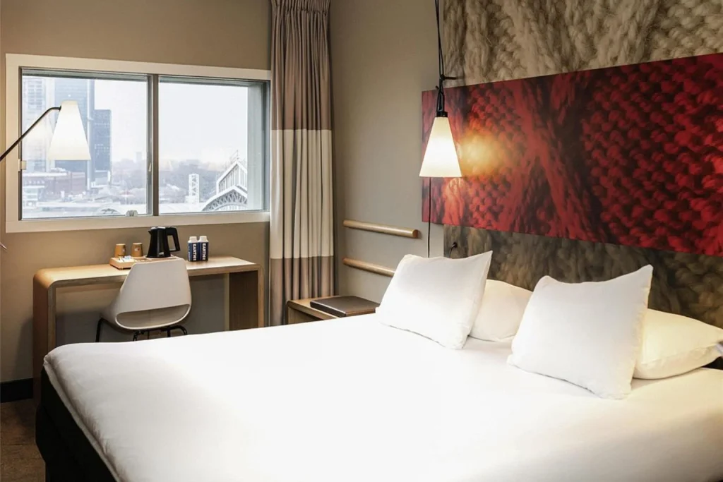 Bright and comfortable double bedroom featuring minimalist furnishings, a practical work desk, stylish red wall art and city views at ibis Amsterdam Centre.