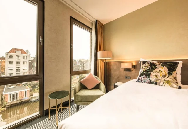 Modern boutique hotel room with comfortable double bed, floral cushions, cozy armchair and large windows overlooking an Amsterdam canal at Monet Garden Hotel Amsterdam