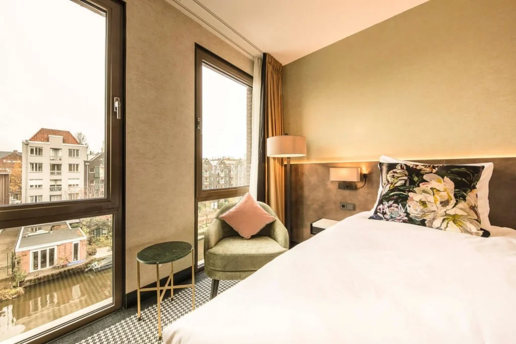 Modern boutique hotel room with comfortable double bed, floral cushions, cozy armchair and large windows overlooking an Amsterdam canal at Monet Garden Hotel Amsterdam