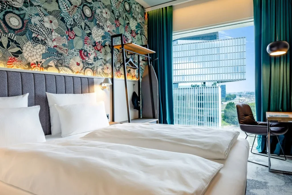 Modern guest room with twin beds, floral feature wall and large city-view window at Motel One Amsterdam, Amsterdam