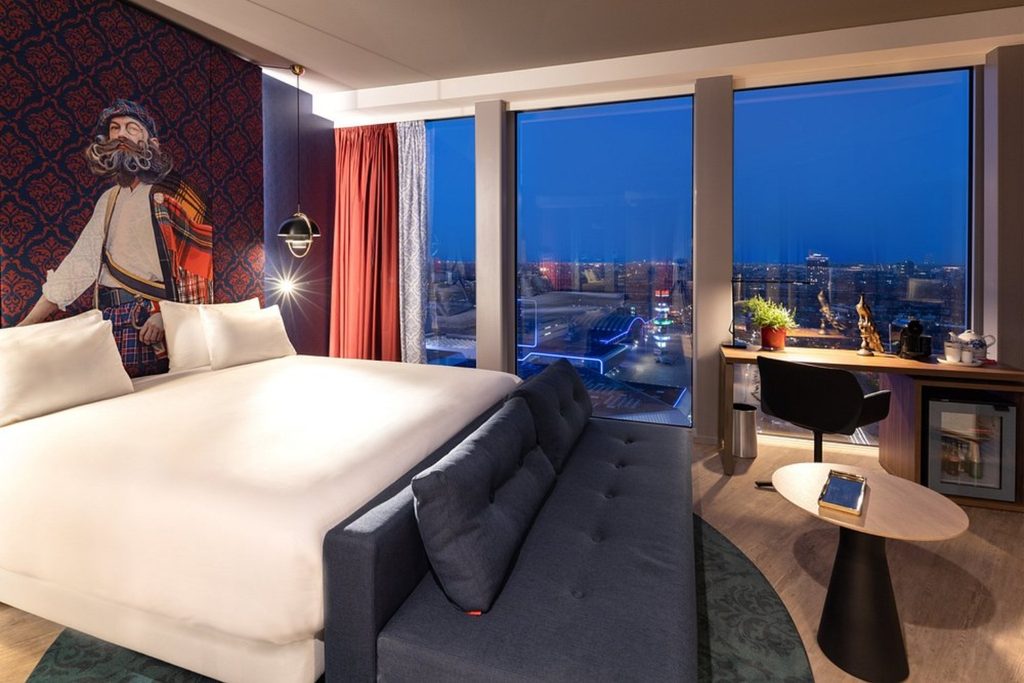 Stylish hotel room with king bed, sofa, work desk and floor-to-ceiling windows with panoramic city views at nhow Amsterdam RAI in Amsterdam