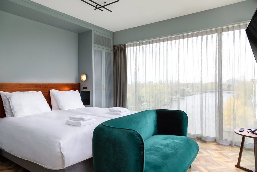 Modern hotel room with double bed, green velvet sofa and floor-to-ceiling windows overlooking the water at Olympic Hotel in Amsterdam