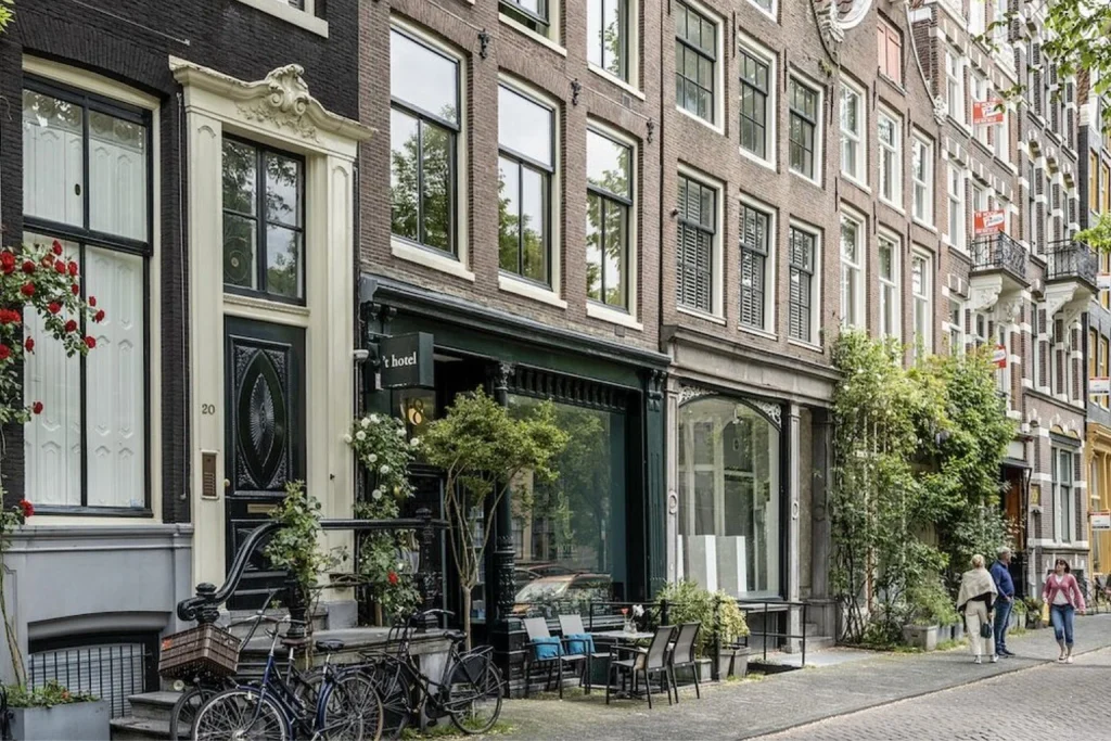Historic canal house facade with tall windows, traditional architecture and a charming tree-lined street setting at ’T Hotel Amsterdam.
