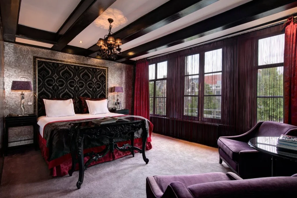 Romantic luxury boutique hotel room with ornate king-size bed, dark wooden ceiling beams, chandelier and purple lounge seating at The Toren in Amsterdam