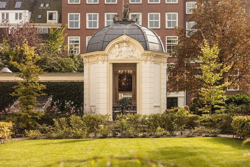 Elegant private garden featuring manicured lawns, mature trees and an ornate historic pavilion surrounded by lush planting, Waldorf Astoria Amsterdam, Amsterdam.